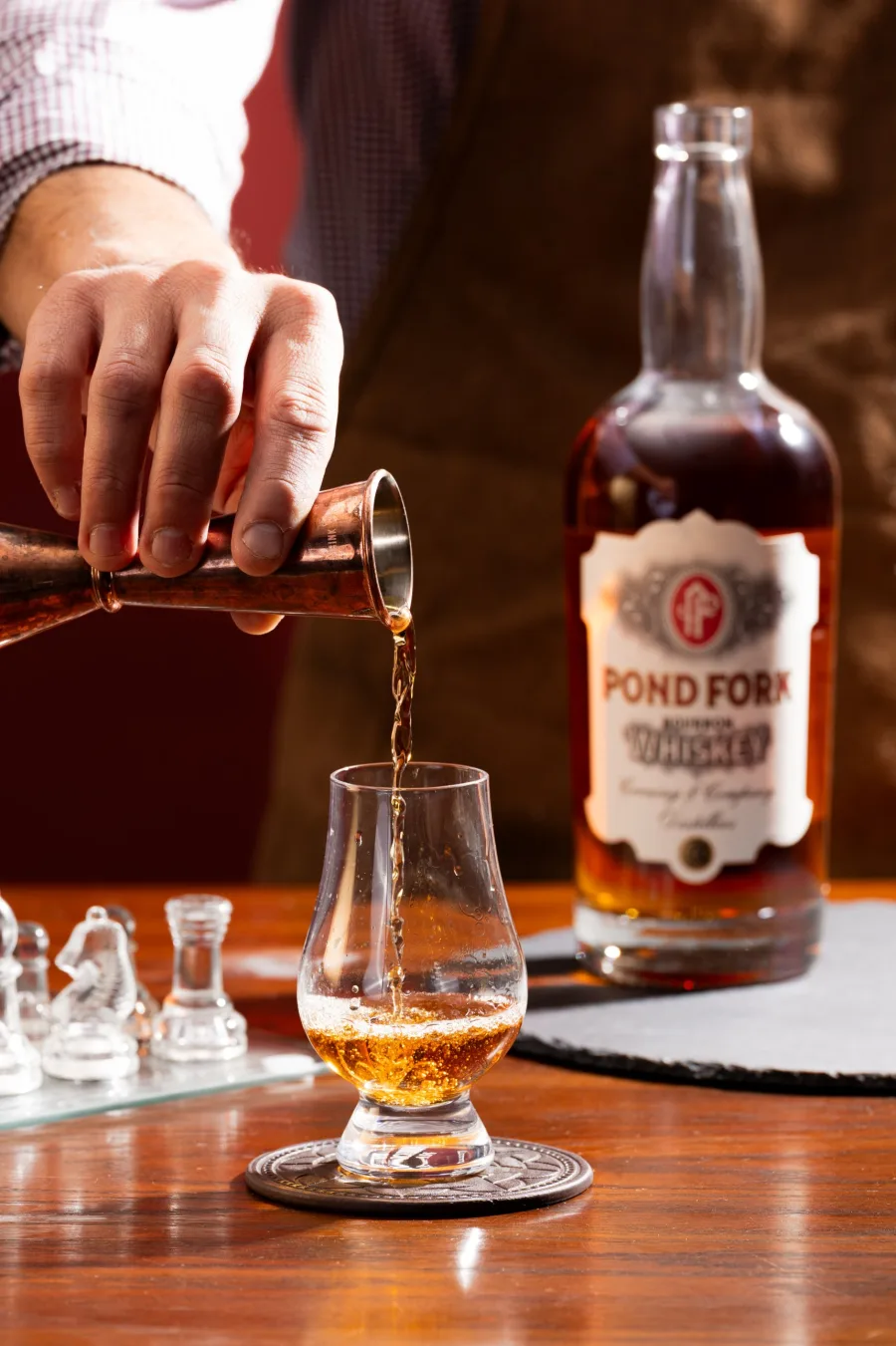 A bartender pours POND FORK Bourbon from a copper jigger into a tasting glass, the bottle beside it on a wooden bar